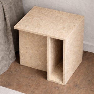 May include: A light brown, cuboid side table with a recessed section. The table has a simple, modern design and is made of a textured material. The table is next to a bed with a gray blanket.