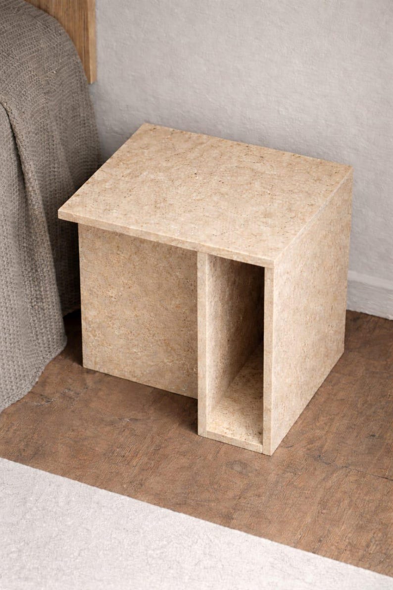 May include: A light brown, cuboid side table with a recessed section. The table has a simple, modern design and is made of a textured material. The table is next to a bed with a gray blanket.