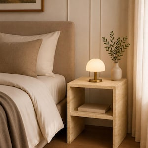 May include: A bedroom scene featuring a beige upholstered bed with pillows and a throw blanket. A small, round lamp with a brass base sits on a wooden nightstand. A vase with greenery is also on the nightstand.
