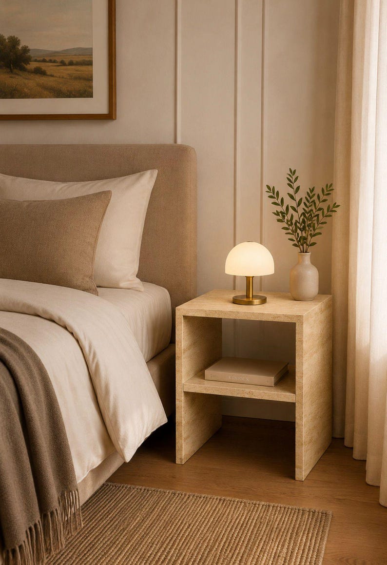 May include: A bedroom scene featuring a beige upholstered bed with pillows and a throw blanket. A small, round lamp with a brass base sits on a wooden nightstand. A vase with greenery is also on the nightstand.