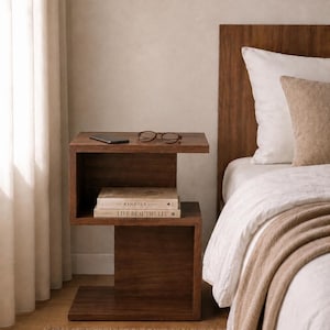 May include: A dark brown wooden nightstand with a modern design. The nightstand has two shelves, with books, a phone, and glasses on top. The bed has white bedding and a brown throw blanket.