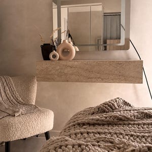 May include: A neutral-toned bedroom scene featuring a modern aesthetic. A large mirror with a glowing frame hangs above a stone shelf holding decorative vases. A cozy chair with a blanket and a chunky knit throw blanket add warmth to the space.