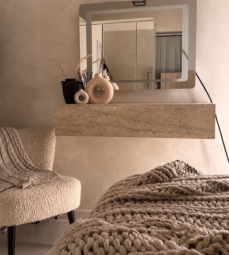 May include: A neutral-toned bedroom scene featuring a modern aesthetic. A large mirror with a glowing frame hangs above a stone shelf holding decorative vases. A cozy chair with a blanket and a chunky knit throw blanket add warmth to the space.