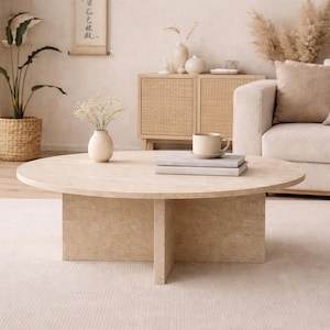 May include: A light beige, round coffee table with a cross-shaped base sits on a light beige rug. A beige sofa with throw pillows is in the background. A woven basket holds a potted plant. A small vase with flowers and a mug are on the table.