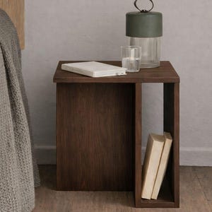 May include: A dark brown wooden nightstand with a rectangular shape. A green lamp, a glass of water, and a closed book sit on top. Two books are visible in the open side. The nightstand is on a wooden floor.