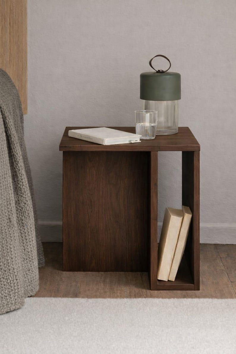 May include: A dark brown wooden nightstand with a rectangular shape. A green lamp, a glass of water, and a closed book sit on top. Two books are visible in the open side. The nightstand is on a wooden floor.