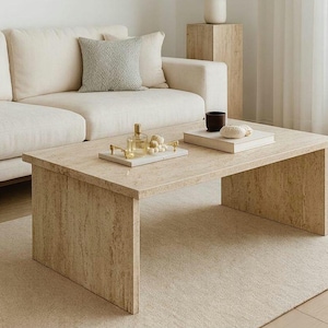 Puede incluir: Una mesa de centro rectangular de piedra beige con una alfombra a juego. Un sofá blanco con un cojín gris está detrás de la mesa. Una bandeja con objetos decorativos y una pequeña taza negra están sobre la mesa.