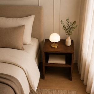 May include: A bedroom scene featuring a bed with beige bedding, a dark wood nightstand, and a small lamp with a white globe shade. A vase with greenery sits on the nightstand. A woven rug is on the floor.
