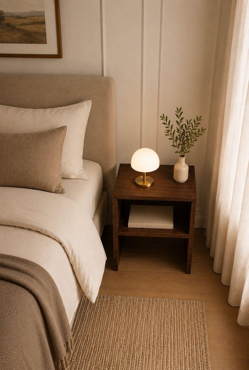 May include: A bedroom scene featuring a bed with beige bedding, a dark wood nightstand, and a small lamp with a white globe shade. A vase with greenery sits on the nightstand. A woven rug is on the floor.