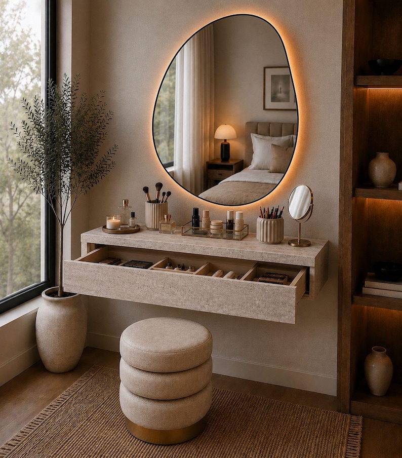 May include: A modern vanity setup with a large, irregularly shaped mirror with a warm, glowing border. The vanity features a floating drawer, a round stool, and various beauty products. A tall plant in a vase sits beside a window.