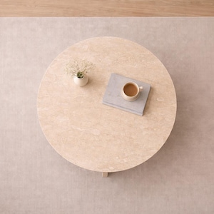May include: Overhead view of a round, light-colored coffee table with a small vase of flowers, a book, and a cup of coffee. The table sits on a neutral-toned rug, with a wooden floor visible on the side.