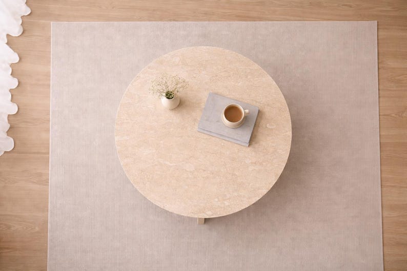 May include: Overhead view of a round, light-colored coffee table with a small vase of flowers, a book, and a cup of coffee. The table sits on a neutral-toned rug, with a wooden floor visible on the side.