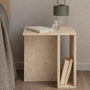 May include: A beige stone side table with a rectangular top and open shelf. A green lamp with a glass base and a glass of water sit on top. Two books are in the shelf. A bed and rug are in the background.