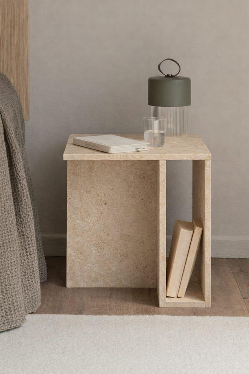 May include: A beige stone side table with a rectangular top and open shelf. A green lamp with a glass base and a glass of water sit on top. Two books are in the shelf. A bed and rug are in the background.