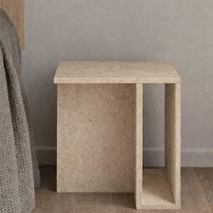 May include: A beige, rectangular side table with a cutout on one side. The table is made of a textured material and sits on a wooden floor. The table is approximately 20 inches tall.