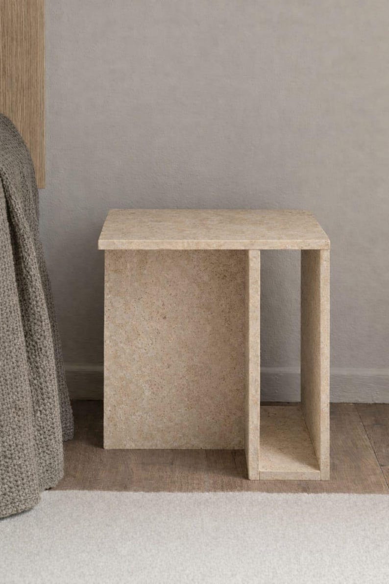 May include: A beige, rectangular side table with a cutout on one side. The table is made of a textured material and sits on a wooden floor. The table is approximately 20 inches tall.