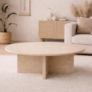 May include: A round, light-colored coffee table with a four-legged base. The table is the focal point of a neutral-toned living room, complemented by a beige rug, sofa, and decorative elements. A potted plant and a cabinet are visible.