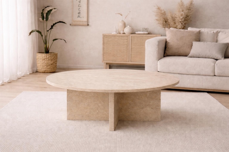 May include: A round, light-colored coffee table with a four-legged base. The table is the focal point of a neutral-toned living room, complemented by a beige rug, sofa, and decorative elements. A potted plant and a cabinet are visible.