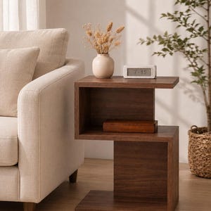 May include: A dark brown wooden side table with a unique, modern design. The table has two shelves and a rectangular shape. A small white clock displaying "7:15" sits on top, next to a vase with dried flowers. A brown box is on the lower shelf.