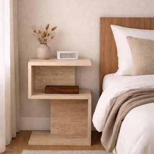 May include: A modern, light-colored wooden nightstand with a unique, geometric design. It features two shelves, a small vase with dried flowers, a digital clock displaying 7:15, and a wooden box. The nightstand is next to a bed with white linens.