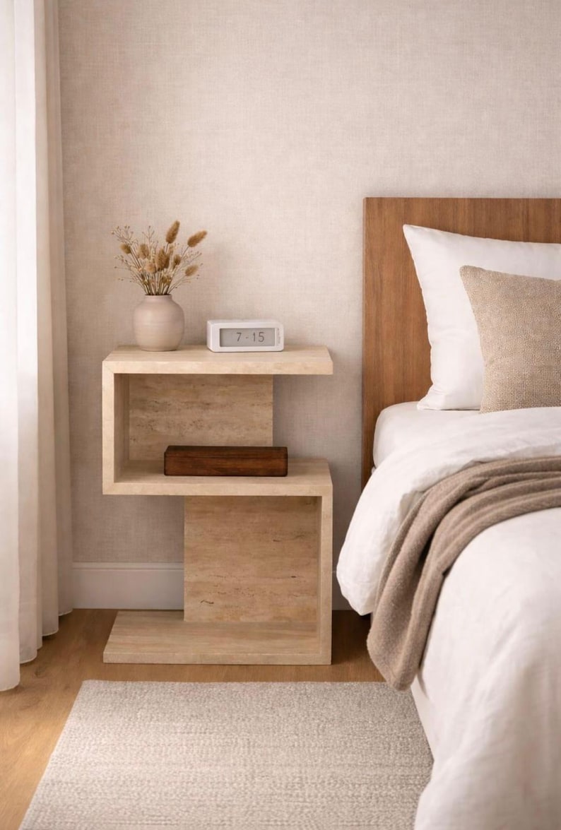 May include: A modern, light-colored wooden nightstand with a unique, geometric design. It features two shelves, a small vase with dried flowers, a digital clock displaying 7:15, and a wooden box. The nightstand is next to a bed with white linens.