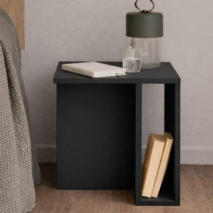 May include: A black bedside table with a rectangular design. A green lamp with a leather handle, a glass of water, and a book rest on top. Two books are visible in the open side of the table.