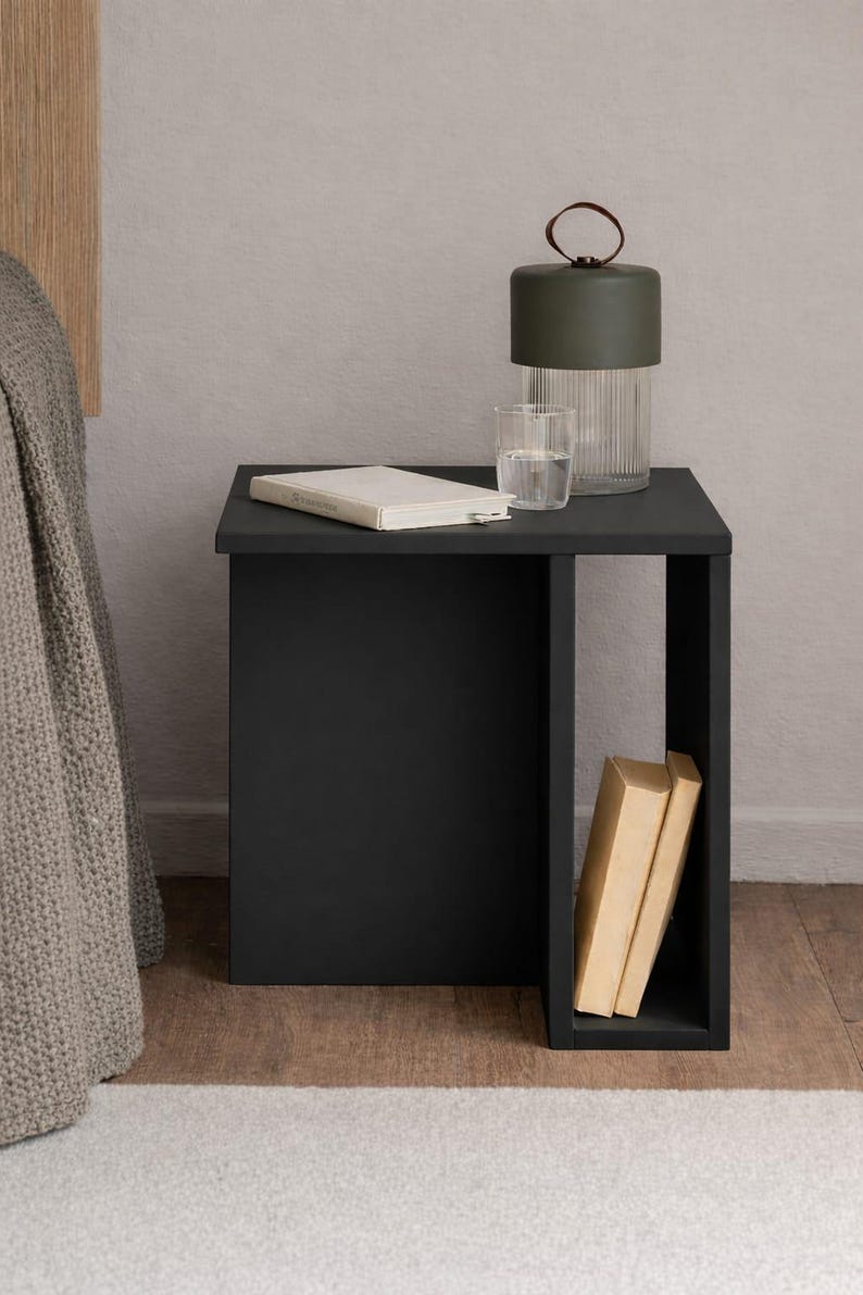 May include: A black bedside table with a rectangular design. A green lamp with a leather handle, a glass of water, and a book rest on top. Two books are visible in the open side of the table.