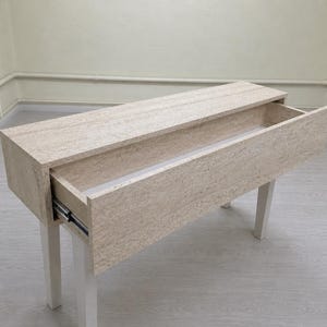 May include: A light-colored rectangular wooden console table with a drawer partially open. The table has four white legs and a light-colored wood grain finish. The drawer slides out on metal tracks.