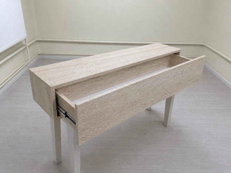 May include: A light-colored rectangular wooden console table with a drawer partially open. The table has four white legs and a light-colored wood grain finish. The drawer slides out on metal tracks.