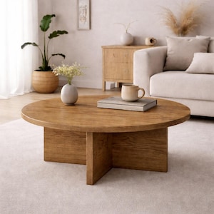 Op de afbeelding: Een ronde houten salontafel met een lichtbruine afwerking. De tafel heeft een kruisvormige basis en is ongeveer 100 cm in diameter. Een boek en een mok staan op de tafel. De tafel staat in een woonkamer.