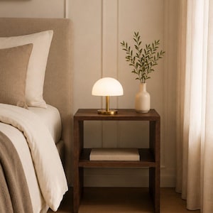 May include: A bedroom scene featuring a dark wood nightstand with a small, white, dome-shaped lamp with a gold base. A white vase with greenery sits next to the lamp. A bed with a beige headboard and bedding is in the background.