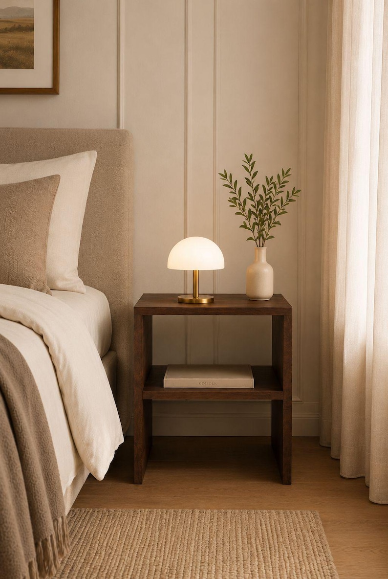 May include: A bedroom scene featuring a dark wood nightstand with a small, white, dome-shaped lamp with a gold base. A white vase with greenery sits next to the lamp. A bed with a beige headboard and bedding is in the background.