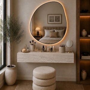 May include: A modern vanity setup with a large, irregularly shaped mirror with a warm, glowing border. The vanity features a stone countertop with makeup and accessories, a three-tiered stool, and a potted plant. The room has a neutral color palette.