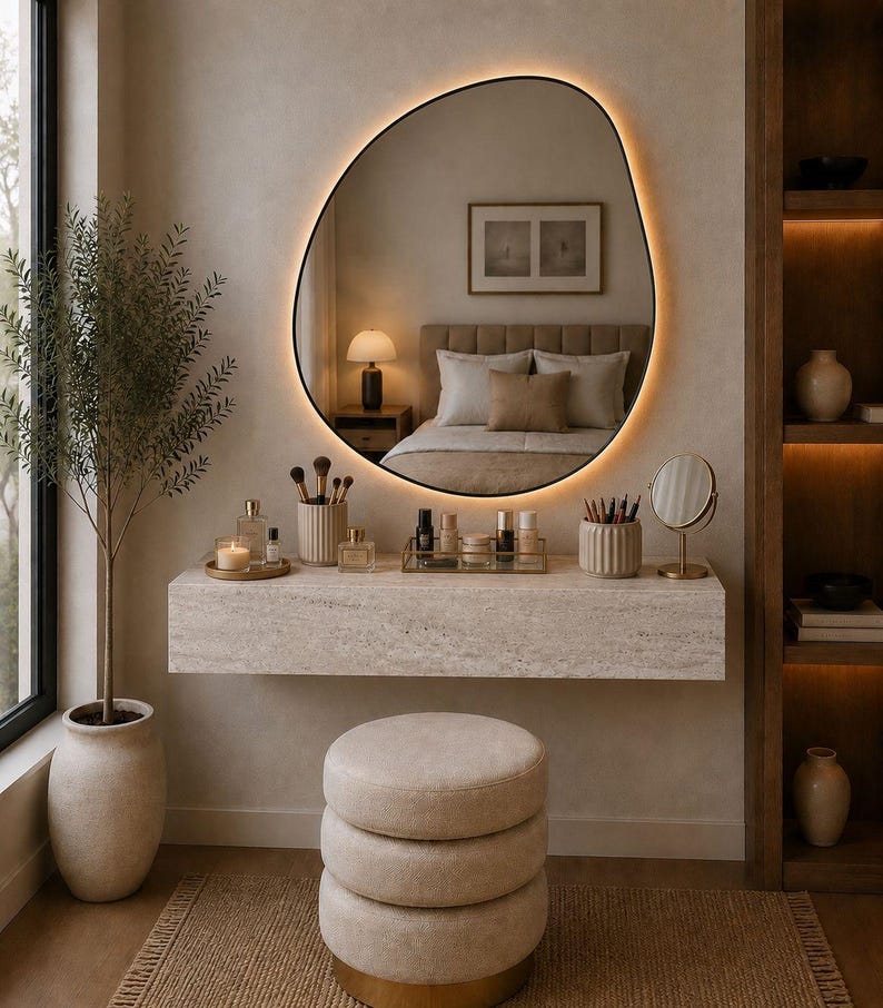 May include: A modern vanity setup with a large, irregularly shaped mirror with a warm, glowing border. The vanity features a stone countertop with makeup and accessories, a three-tiered stool, and a potted plant. The room has a neutral color palette.