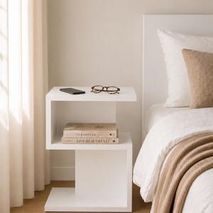 May include: A modern white bedside table with a unique, open design. The table holds a smartphone, eyeglasses, and two books titled "THE KINFOLK HOME" and "LIVE BEAUTIFULLY". The scene includes a bed with white linens and a tan throw blanket.