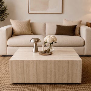 Pode incluir: Sala de estar em tons neutros com um sofá bege, almofadas e uma mesa de centro quadrada de pedra. Uma lâmpada prateada e um vaso com flores brancas estão sobre a mesa. Um tapete tecido e um móvel de madeira completam a cena. A mesa tem cerca de 100 cm.