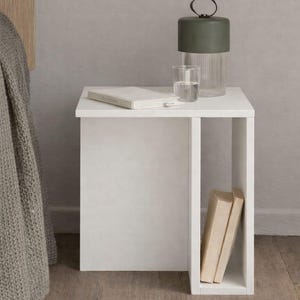 May include: A white bedside table with a built-in shelf holds a green lamp, a glass of water, and a book. Two books are stored in the shelf. The table is next to a bed with a gray blanket.
