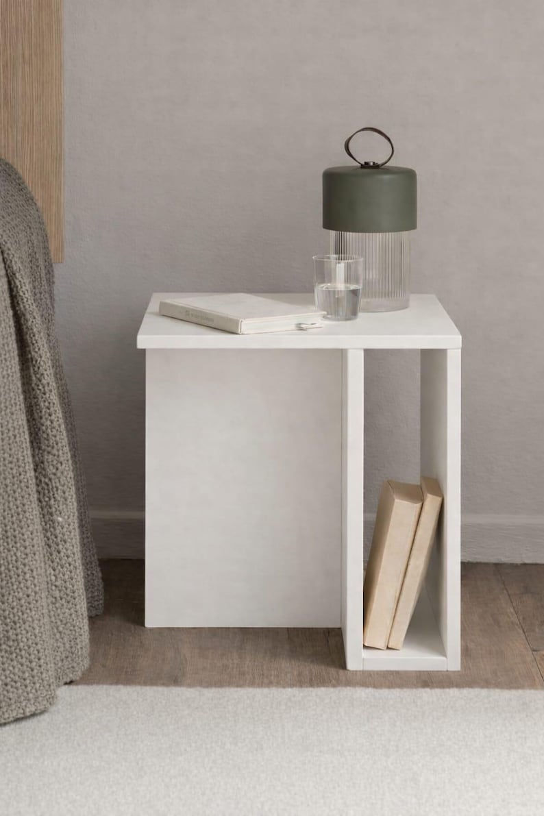 May include: A white bedside table with a built-in shelf holds a green lamp, a glass of water, and a book. Two books are stored in the shelf. The table is next to a bed with a gray blanket.