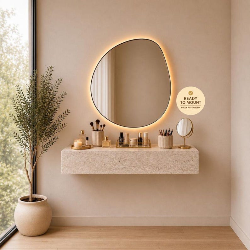 May include: A modern vanity setup featuring a large, irregularly shaped mirror with a warm, glowing border. The mirror hangs above a stone-textured floating shelf holding makeup and accessories. A potted plant and a small round mirror complete the elegant design.