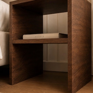 May include: A dark brown wooden nightstand with a shelf holding a book labeled "KINFOLK". The nightstand has a minimalist design and is placed next to a bed. A lamp and vase are on top.