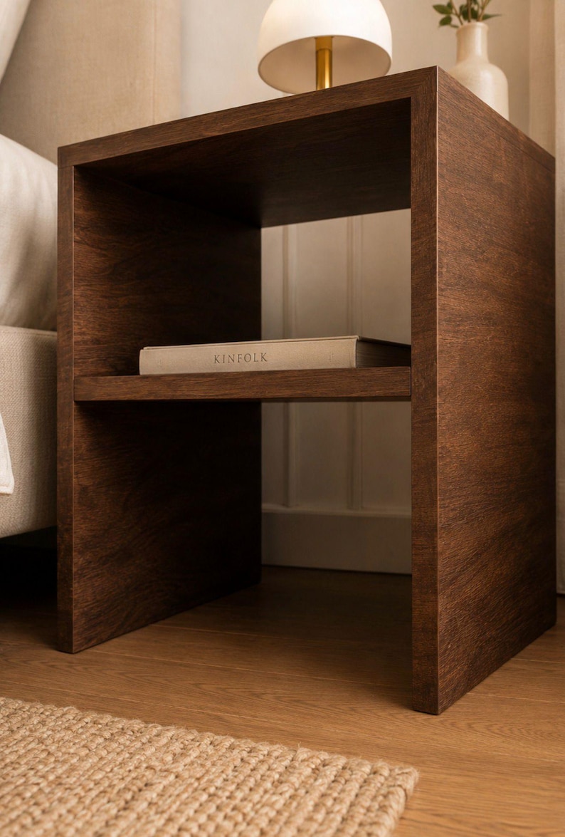 May include: A dark brown wooden nightstand with a shelf holding a book labeled "KINFOLK". The nightstand has a minimalist design and is placed next to a bed. A lamp and vase are on top.