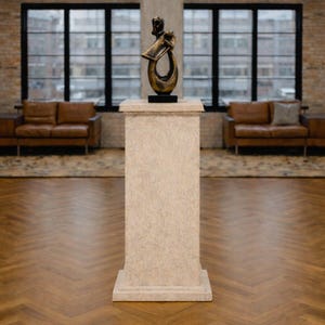 K&ouml;nnte beinhalten: Eine Bronzeskulptur auf einem Steinsockel. Die Skulptur hat ein abstraktes Design mit dunkler Basis. Der Sockel ist rechteckig und strukturiert. Der Hintergrund umfasst Fenster, Backsteinmauern und Ledersofas.