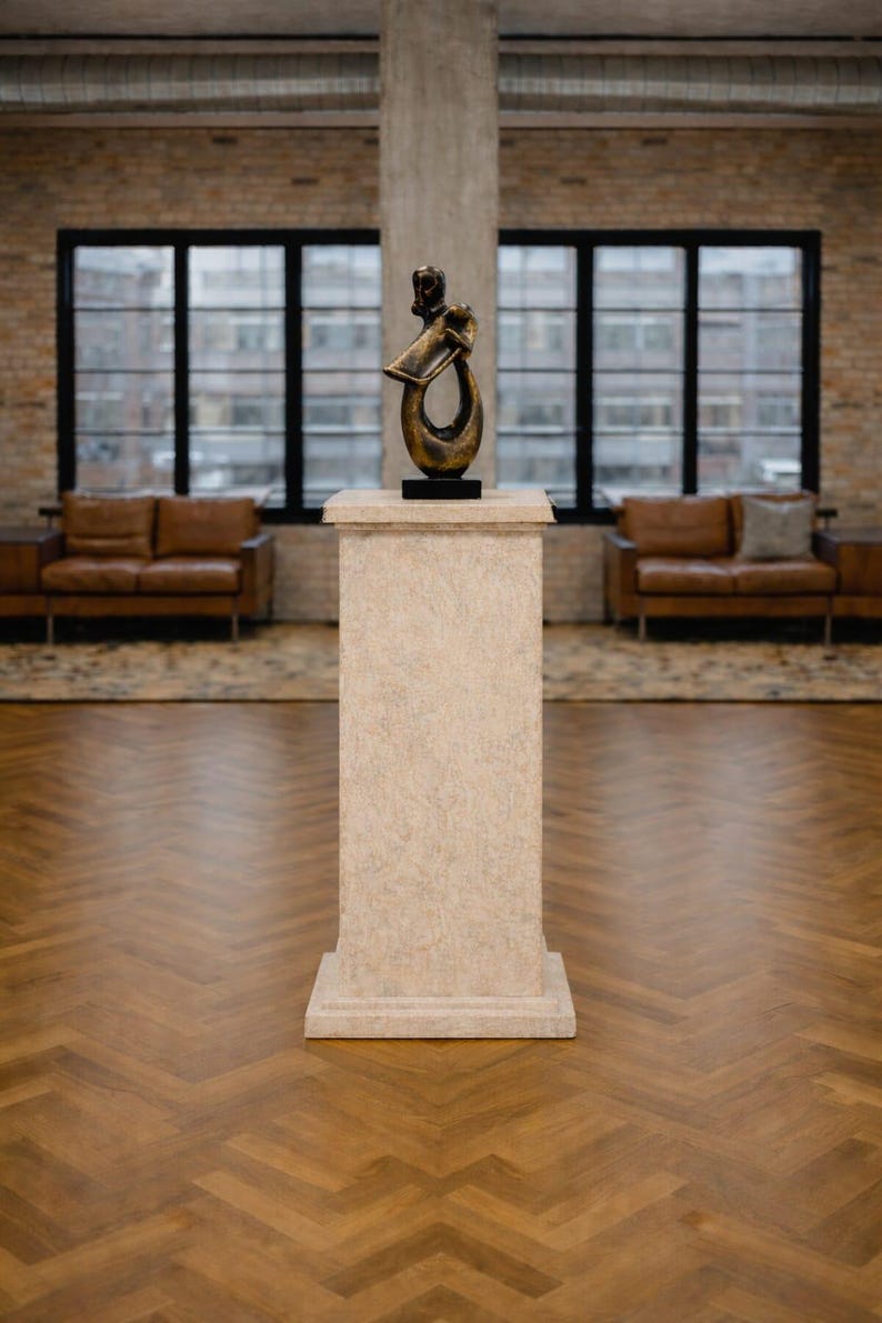 K&ouml;nnte beinhalten: Eine Bronzeskulptur auf einem Steinsockel. Die Skulptur hat ein abstraktes Design mit dunkler Basis. Der Sockel ist rechteckig und strukturiert. Der Hintergrund umfasst Fenster, Backsteinmauern und Ledersofas.