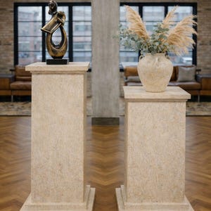 K&ouml;nnte beinhalten: Zwei beige Sockel mit strukturierter Oberfl&auml;che stehen auf einem Holzboden. Ein Sockel tr&auml;gt eine Bronzeskulptur, der andere eine beige Vase mit getrockneten Pflanzen. Der Raum hat gro&szlig;e Fenster und eine Backsteinmauer.
