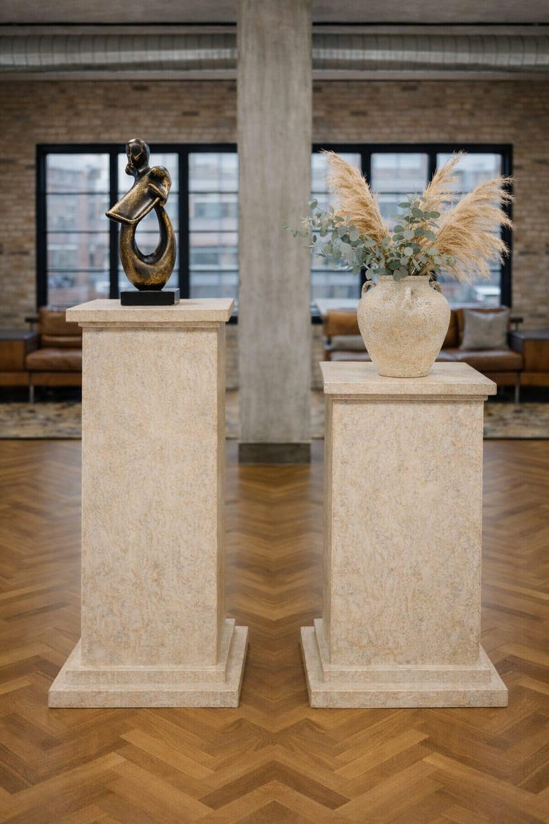 K&ouml;nnte beinhalten: Zwei beige Sockel mit strukturierter Oberfl&auml;che stehen auf einem Holzboden. Ein Sockel tr&auml;gt eine Bronzeskulptur, der andere eine beige Vase mit getrockneten Pflanzen. Der Raum hat gro&szlig;e Fenster und eine Backsteinmauer.