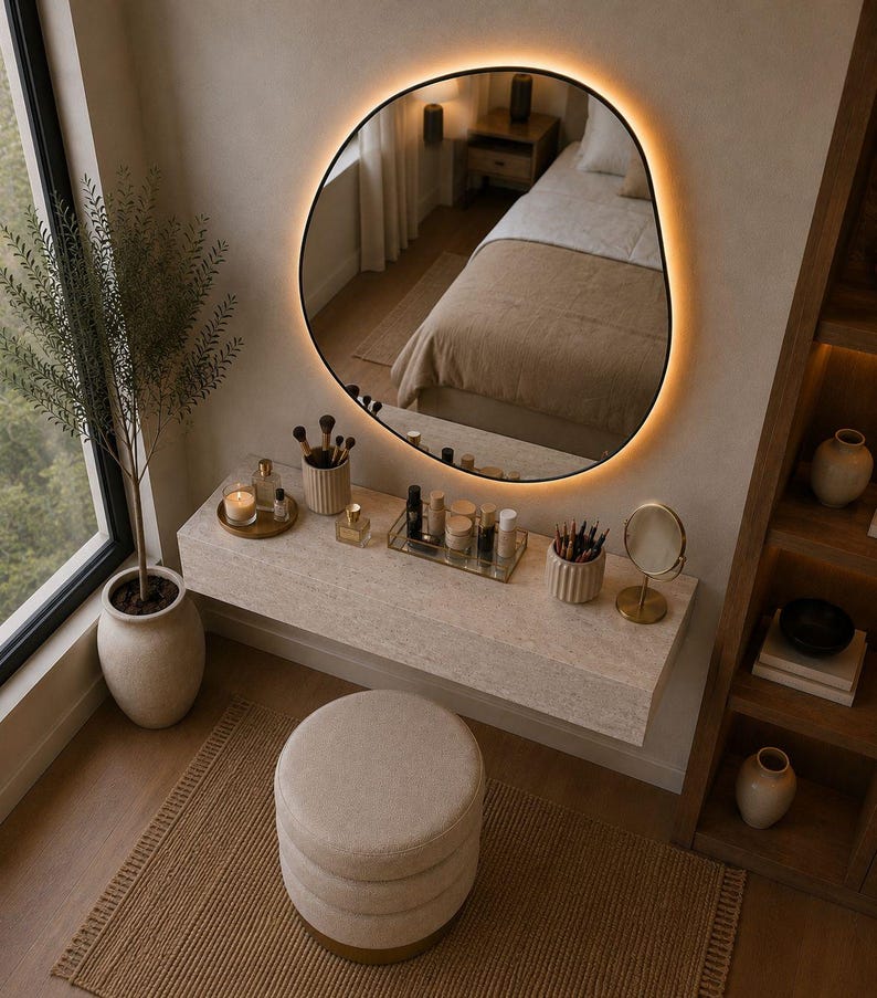 May include: A modern vanity setup with a large, irregularly shaped mirror with a warm, glowing border. The vanity features a light-colored countertop with makeup and brushes, a round stool, and a decorative rug. A potted plant adds a touch of nature.