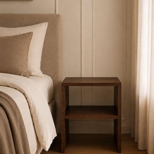 May include: A dark brown wooden nightstand with two shelves stands next to a bed with a beige headboard and bedding. A woven rug is on the floor. The room has a neutral color palette with a painting on the wall.