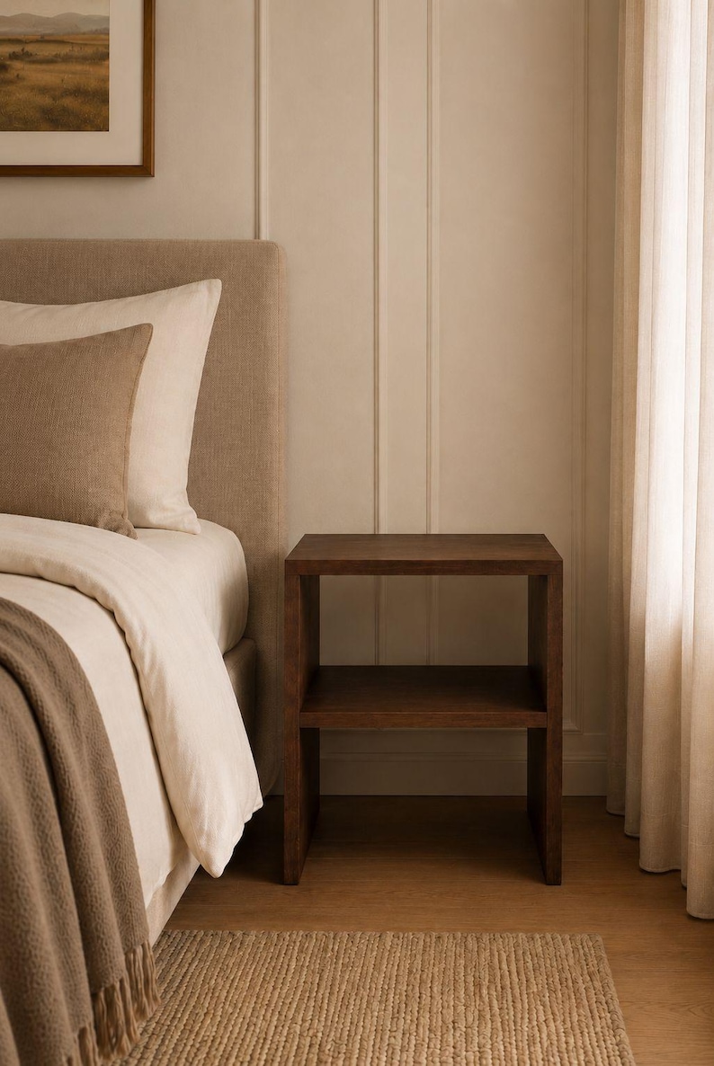 May include: A dark brown wooden nightstand with two shelves stands next to a bed with a beige headboard and bedding. A woven rug is on the floor. The room has a neutral color palette with a painting on the wall.
