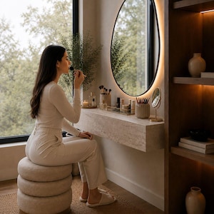 May include: A woman applying makeup at a vanity. The vanity features an oval mirror with a black frame and a built-in light. The countertop holds makeup brushes, perfumes, and other beauty products. A stack of round, beige stools provides seating.