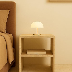 May include: A modern bedroom scene featuring a small, white, dome-shaped table lamp with a gold base on a light wood nightstand. A book is on the lower shelf. The bed and wall are a matching beige color.
